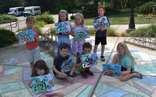 Seven smiling children showing off their artwork outside of the Community Center on a sunny day.